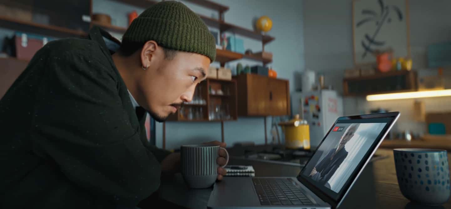 Man in beanie hat with coffee watching live stream of executive woman on laptop