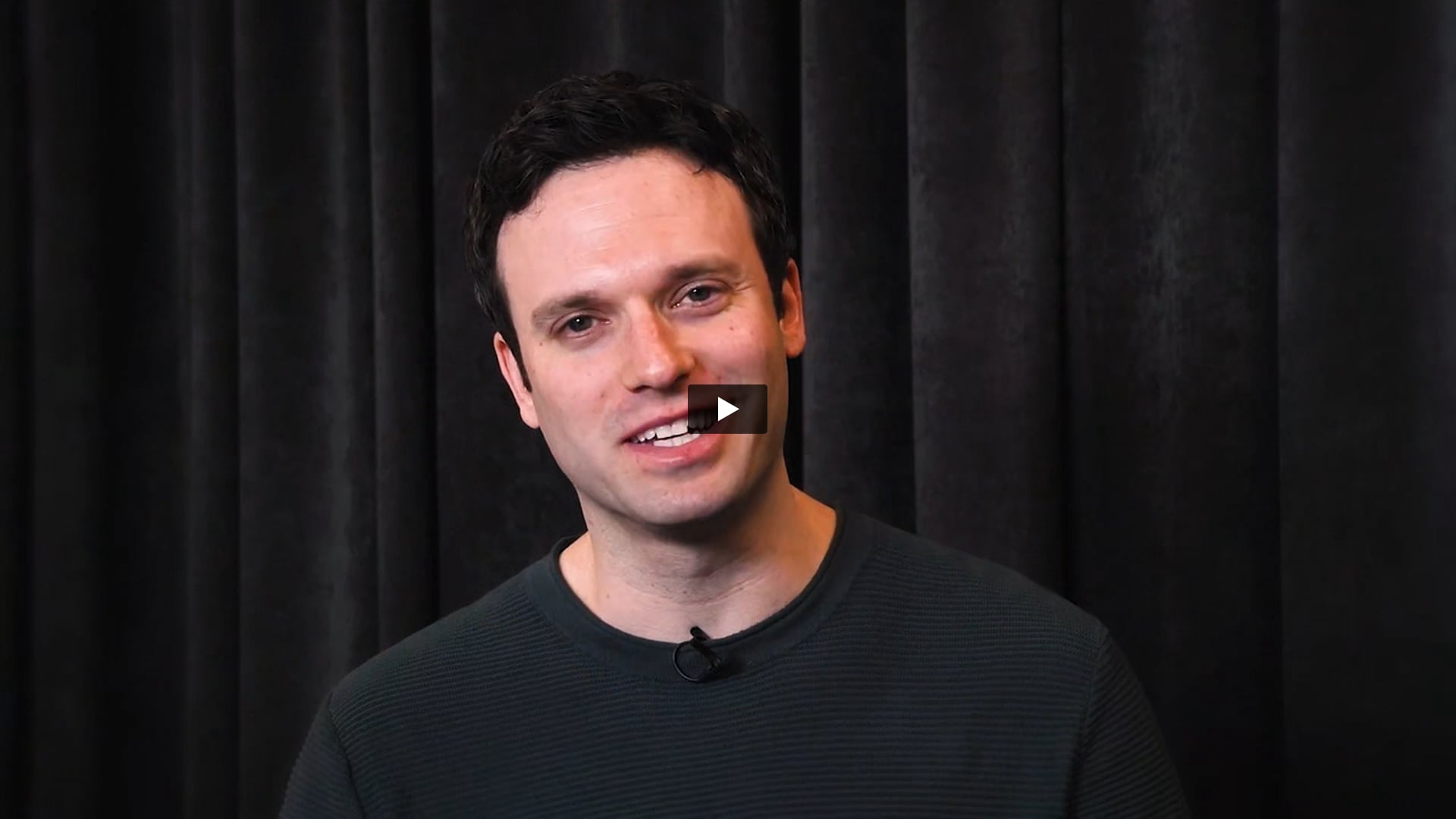 Man with short dark hair and grey shirt standing in front black curtains