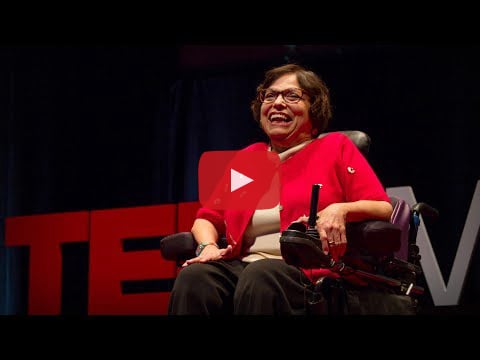 Judy Heumann sitting in her motorized wheelchair, smiling in a red sweater on top of a tan sweater, black pants, and glasses, in front of a red "TED Mid Atlantic" sign and a black curtain.