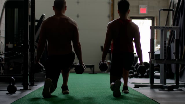 A pair of young fit men do weighted lunges in tandem. 