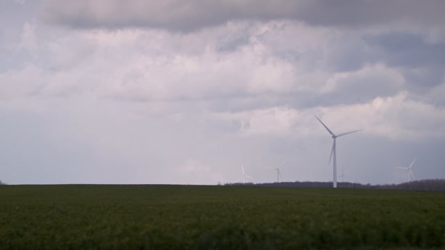 Skies full of clouds float above a fleet of green energy windmills standing in a rural field.