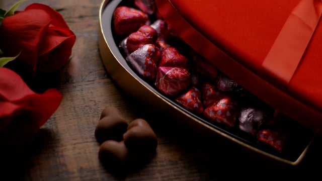 A heart-shaped box of chocolates in wrappers on wooden table with roses. The light slowly fades up. 