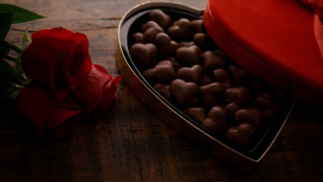 Heart shaped box of chocolates in wrappers on black surface with roses. Light fades in and out. 