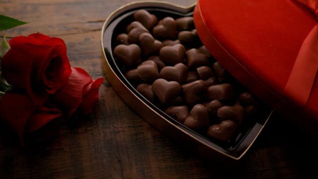 A heart-shaped box of chocolates on a wooden table with roses with camera movement.
