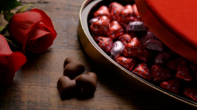 A heart-shaped box of chocolates on a wooden table with roses with camera movement.