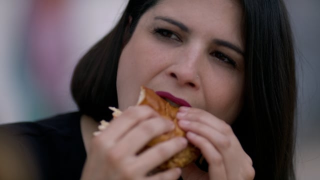 A young woman takes a big satisfying bite of a fresh boutique hamburger with all the fixings. 