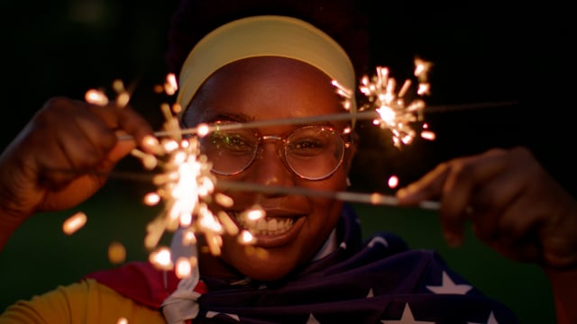 A black woman smiles draped in the flag-waving sparklers. 