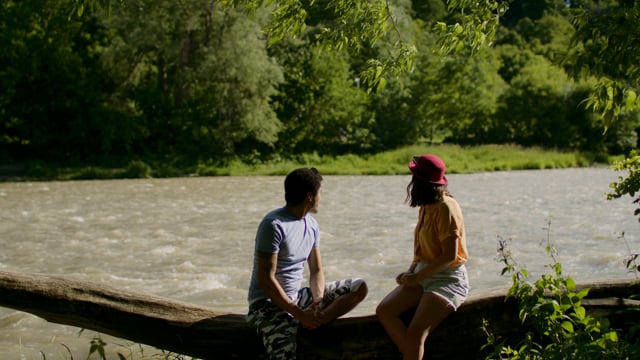 An authentic diverse couple has a romantic day in a park. Sitting on a log with a picturesque river running behind the. Young love.