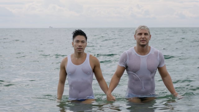An LGBTQ couple holds each other and embraces in a romantic display of love as they exit the crystal clear waters at the beach on holiday. 