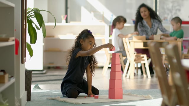 A young student plays in a bright modern classroom while teacher interacts with other children in background. 