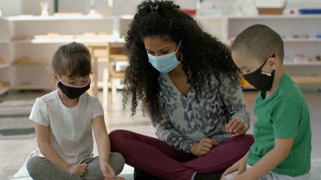 Young teacher and young students engage in a lesson while wearing masks in bright, modern classroom. 