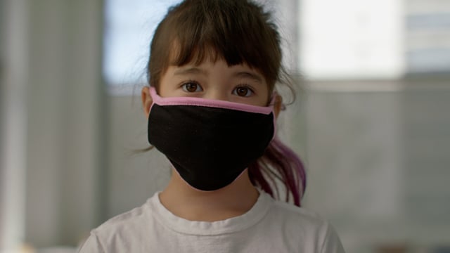Adorable young girl with mask stands smiling in the classroom of a bright modern school.