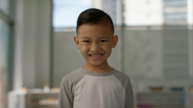 Adorable young boy stands smiling in classroom of bright modern school.