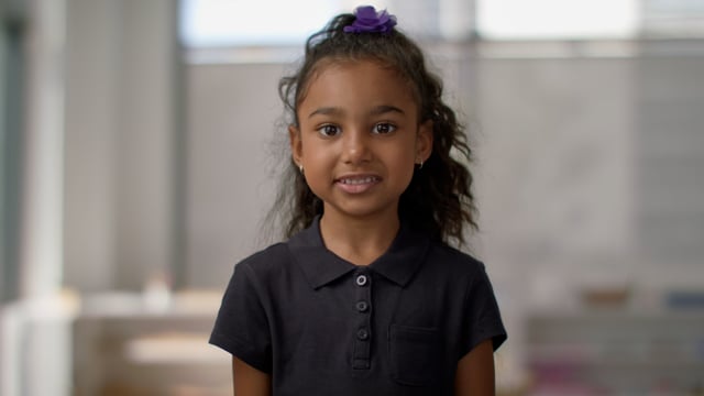 An adorable young girl stands smiling in a modern classroom.