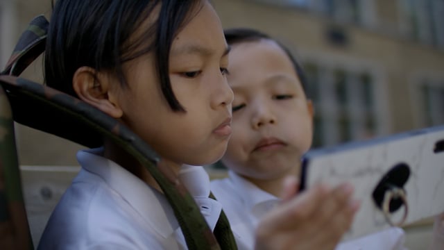 Two young brothers watch an educational video on a smartphone outside their school on their first day back. 