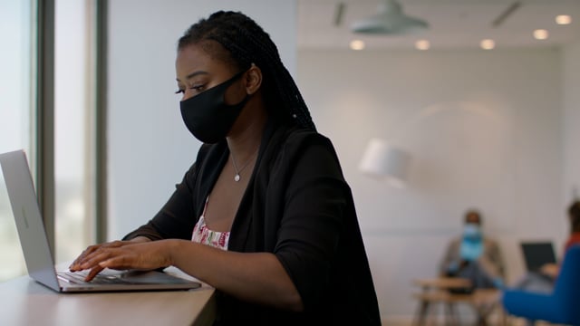 Back at work with social distance precautions. A black woman at a tech firm works on her laptop. Business is back.