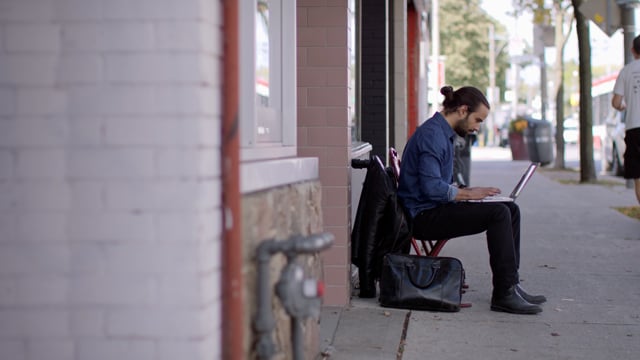 A young man outside of a cafe attentively working remotely on his laptop. 
