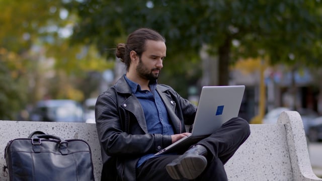 A young man sitting in the park attentively working remotely on his laptop outside of a cafe. 