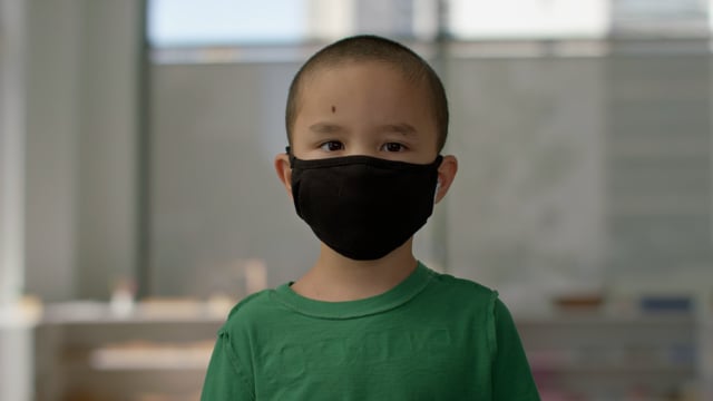 Adorable young boy with mask on stands smiling in classroom of bright modern school.