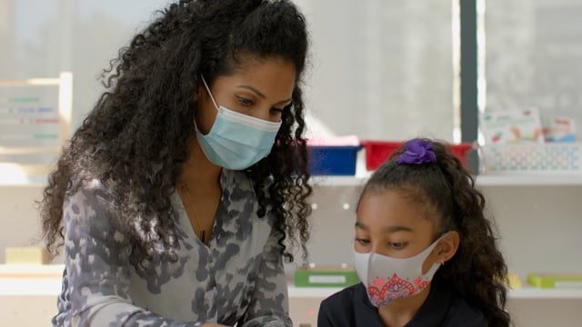 Young female teacher and student engage in lesson while wearing masks in bright, modern classroom. 