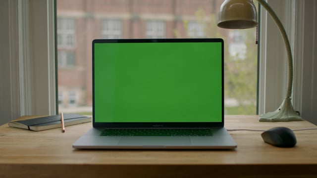 Green screen laptop computer sitting on a home work desk next to a desk lamp.