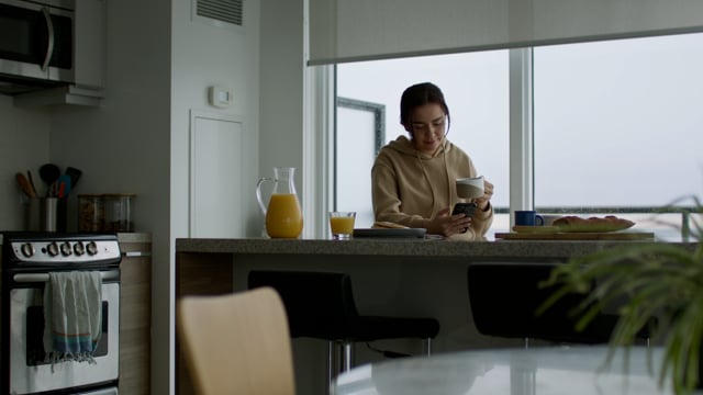 Young woman chilling in her condo, having brunch, checking her phone. 