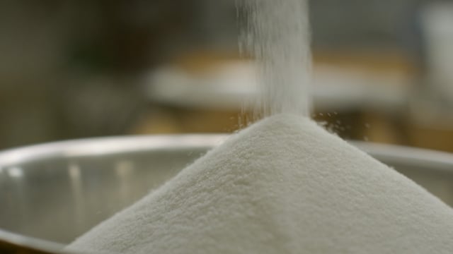 Granulated white sugar pours hypnotically into a large mixing bowl. 