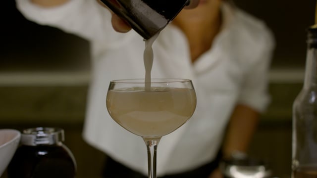 A perfectly executed cocktail is poured into a beautiful coupe glass by a young female bartender. 