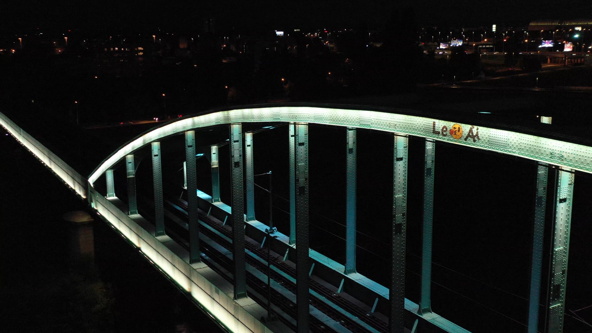 Night view of the lit up light blue train bridge over the river, city lights in the background, graffiti on the bridge