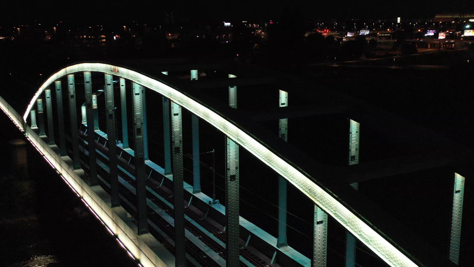 Night view of the lit up light blue train bridge over the river, city lights in the background, rotating