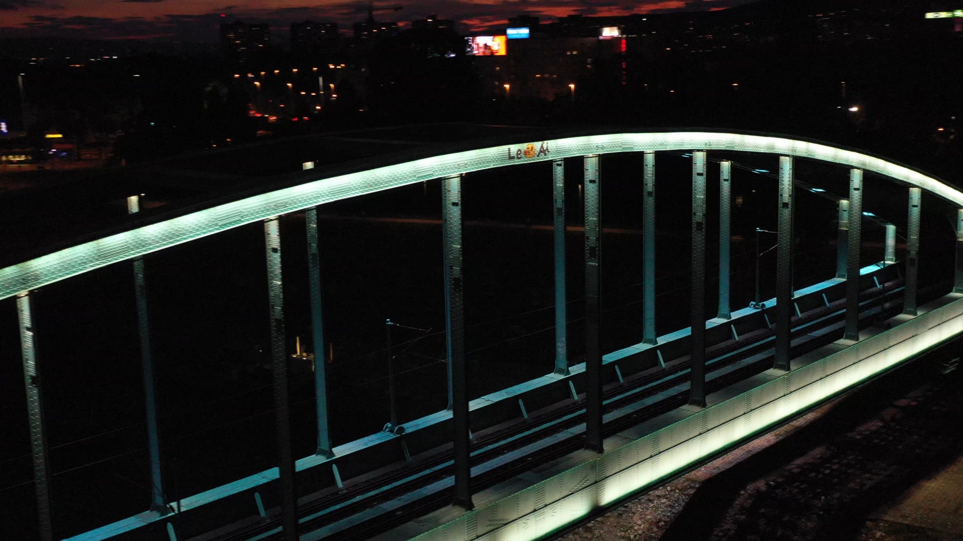 Night view of the lit up light blue train bridge over the river, city lights in the background