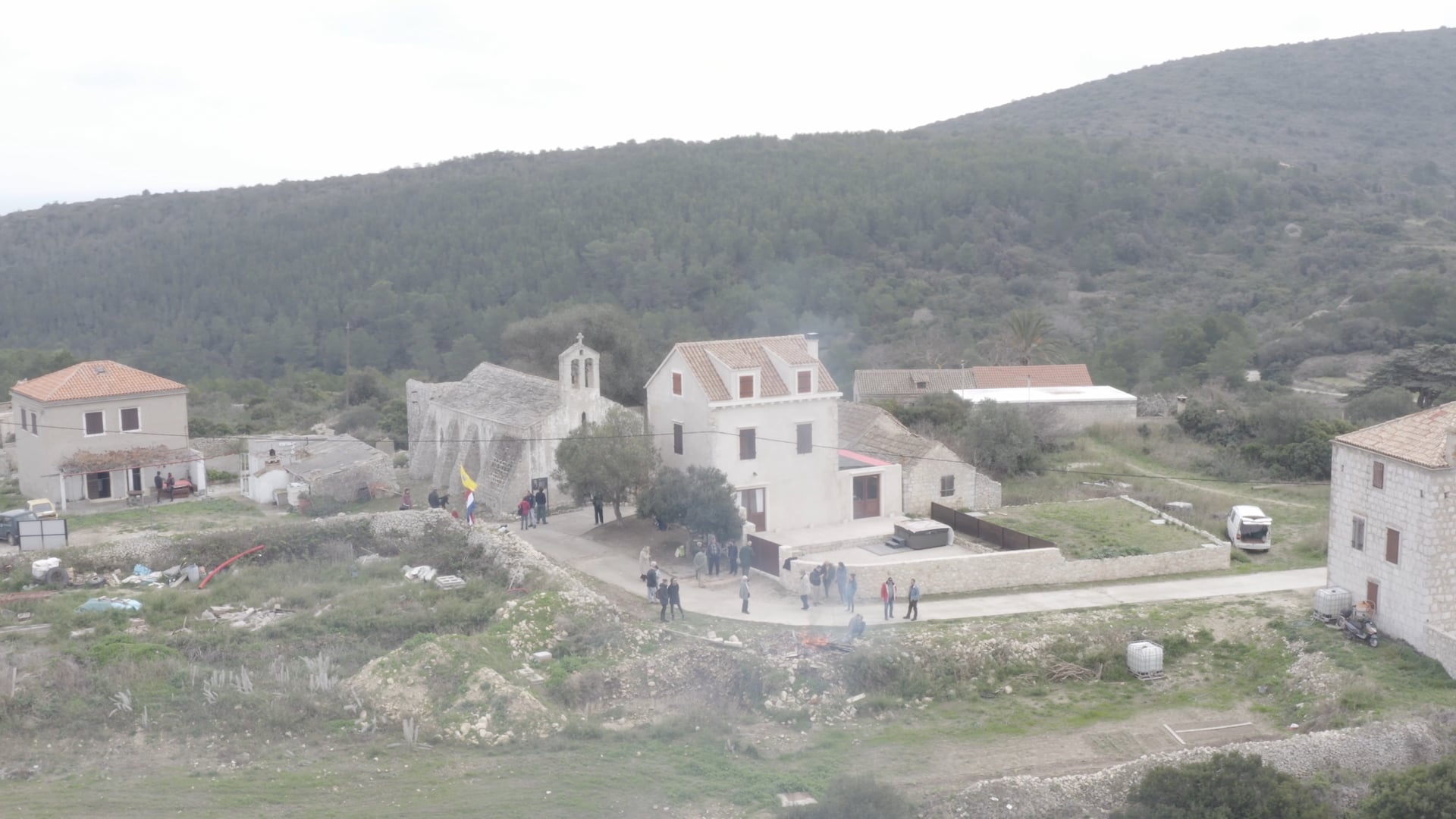 People walking on the road by the church, flags waving, few houses around, 360° view, old white van, small village