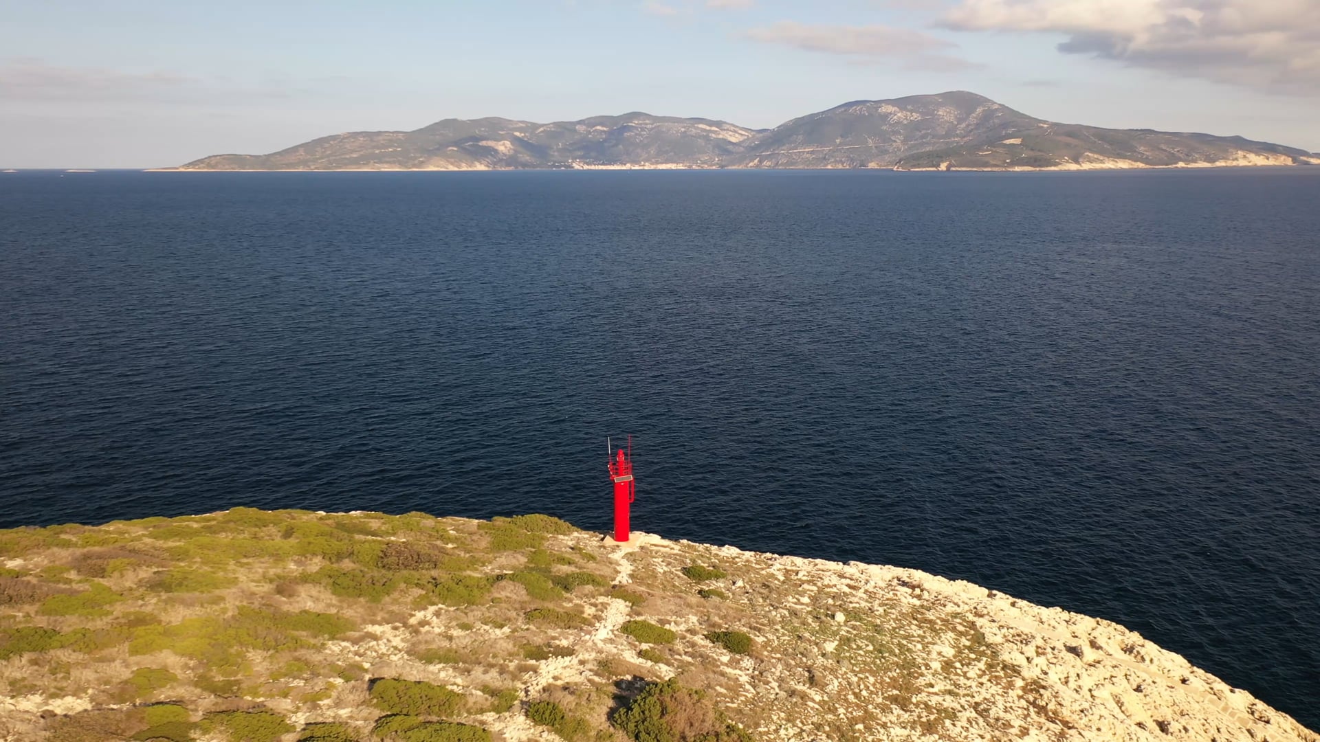 Small red lighthouse on the stone cliff, deep blue, calm sea, other island visible in the distance across the sea