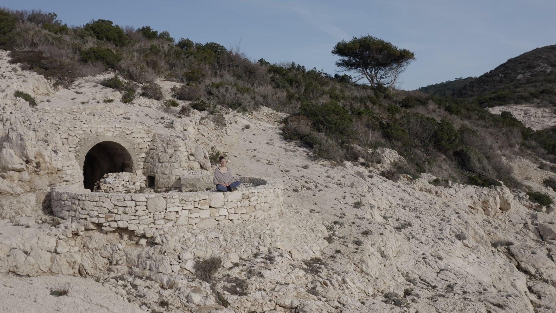 Woman sitting peacefully on the stone wall, slowly moving away, few bushes and a tree in the background