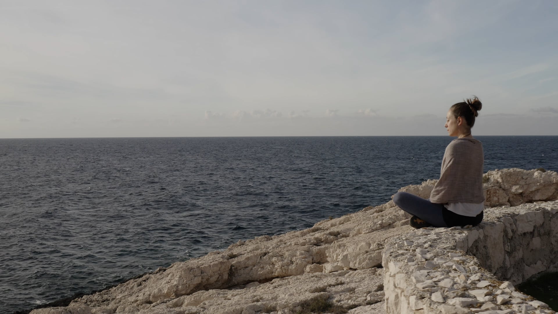 Woman streching, doing yoga, sitting peacefully on the stone wall, looking at the waves and the sun, windy weather