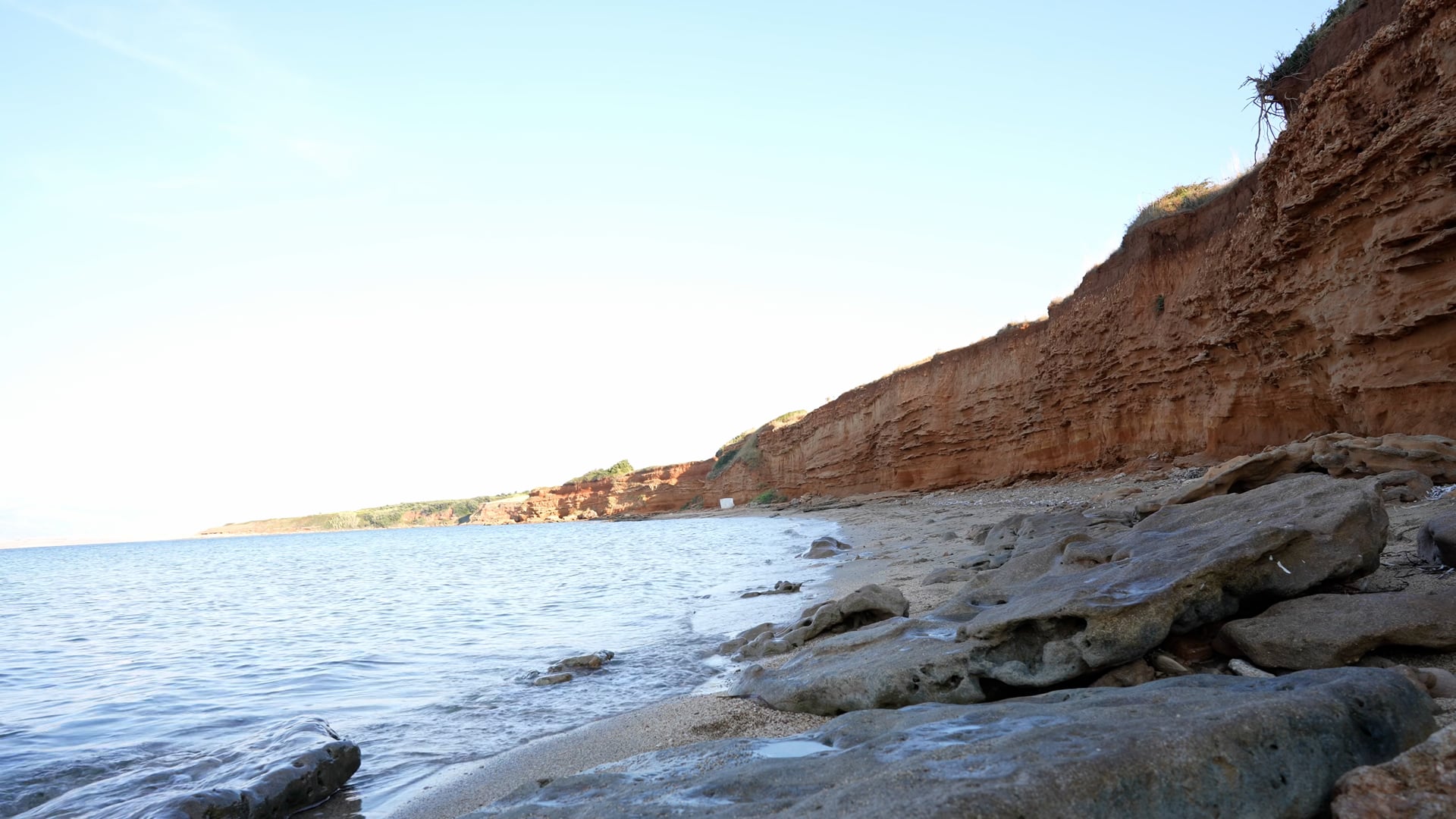 Waves lightly splashing the coast, steady shot, red rocks near the sand beach, sunshine, shadows