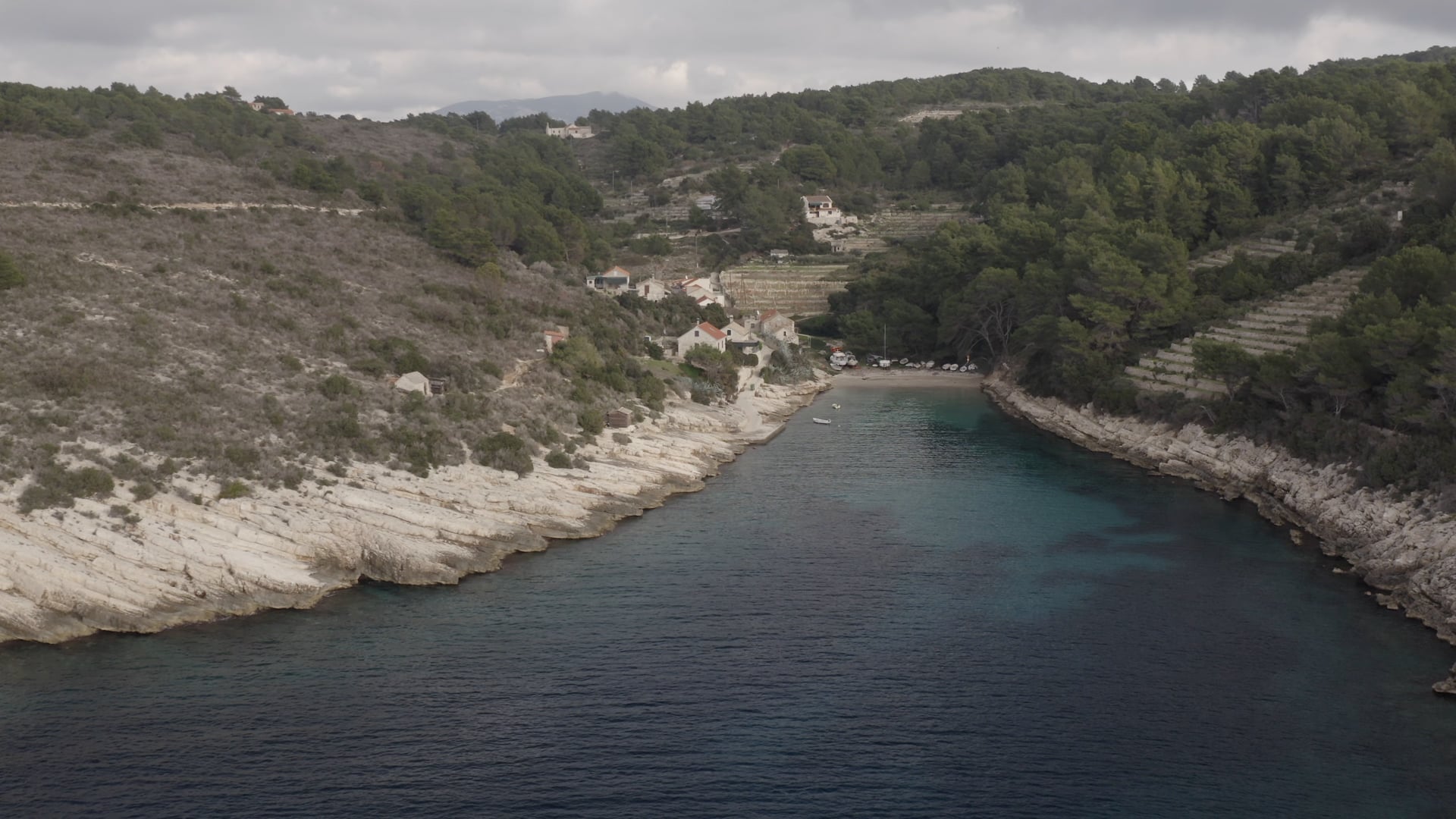 A lot of small boats docked in the small bay, few stone houses and forest behind