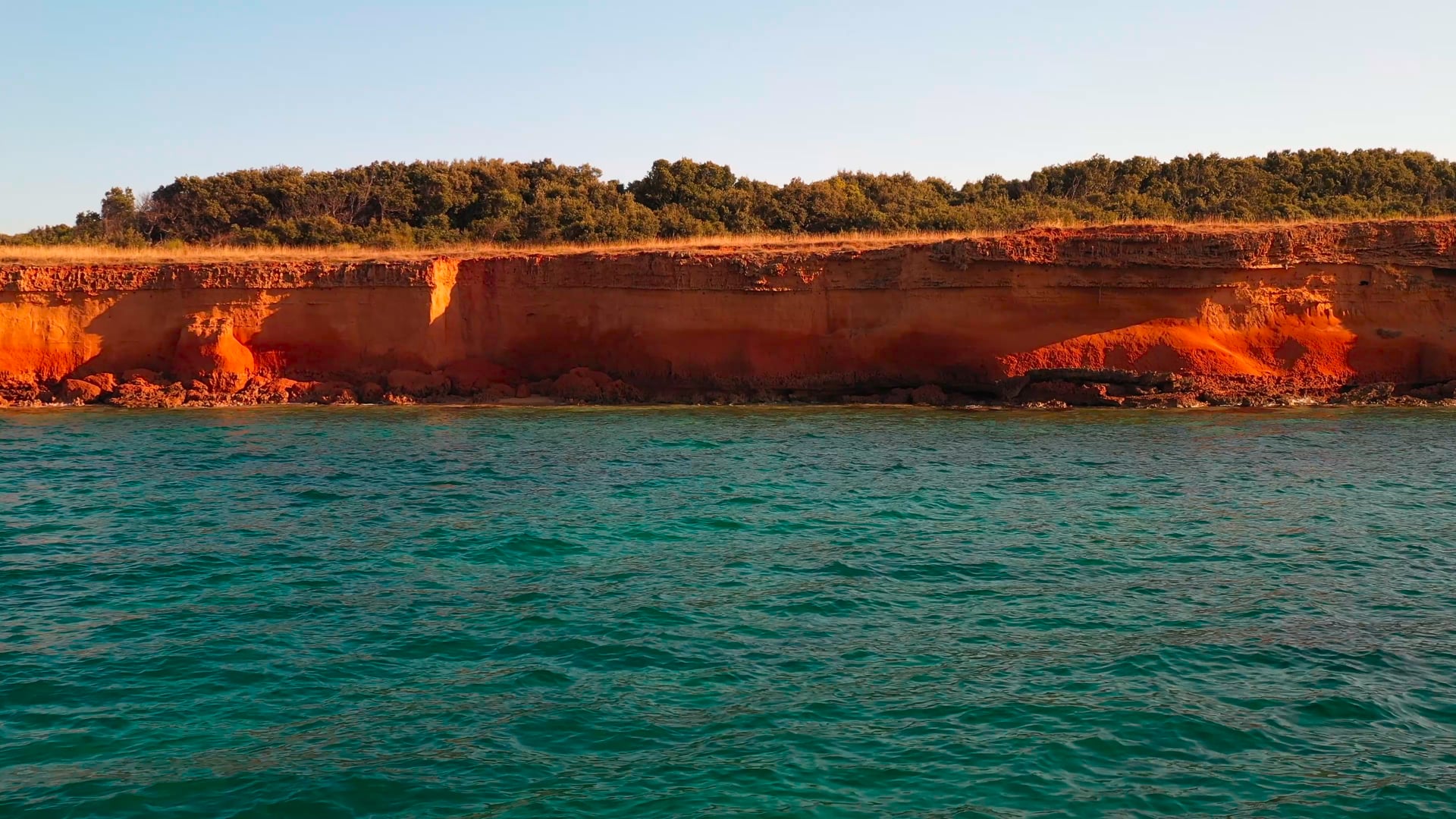 Slowly approaching red stone cliff near the light blue sea, forest in the background, panning back to the sea
