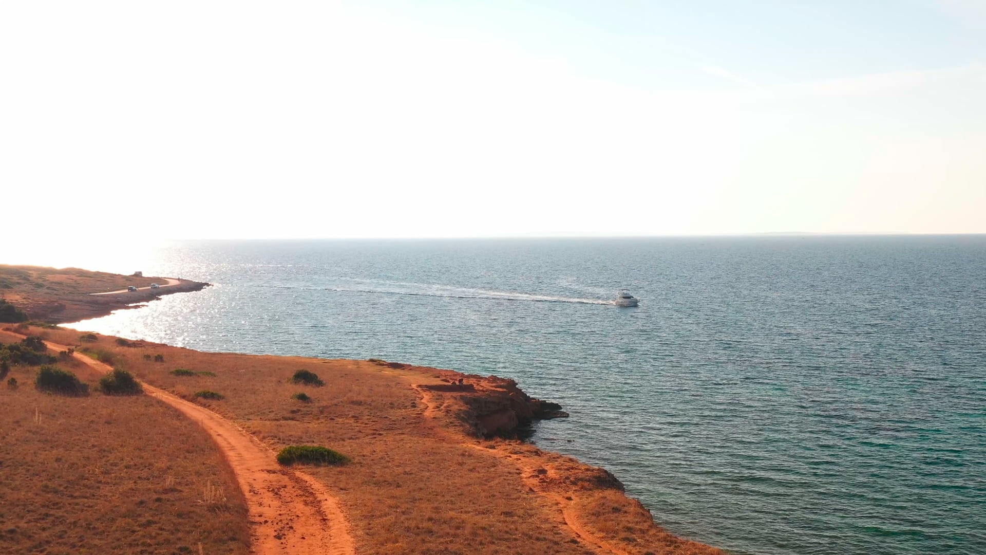 Dirt road on the island, small yacht slowly sailing, crystal clear blue sea, island visible in the distance, extended shot