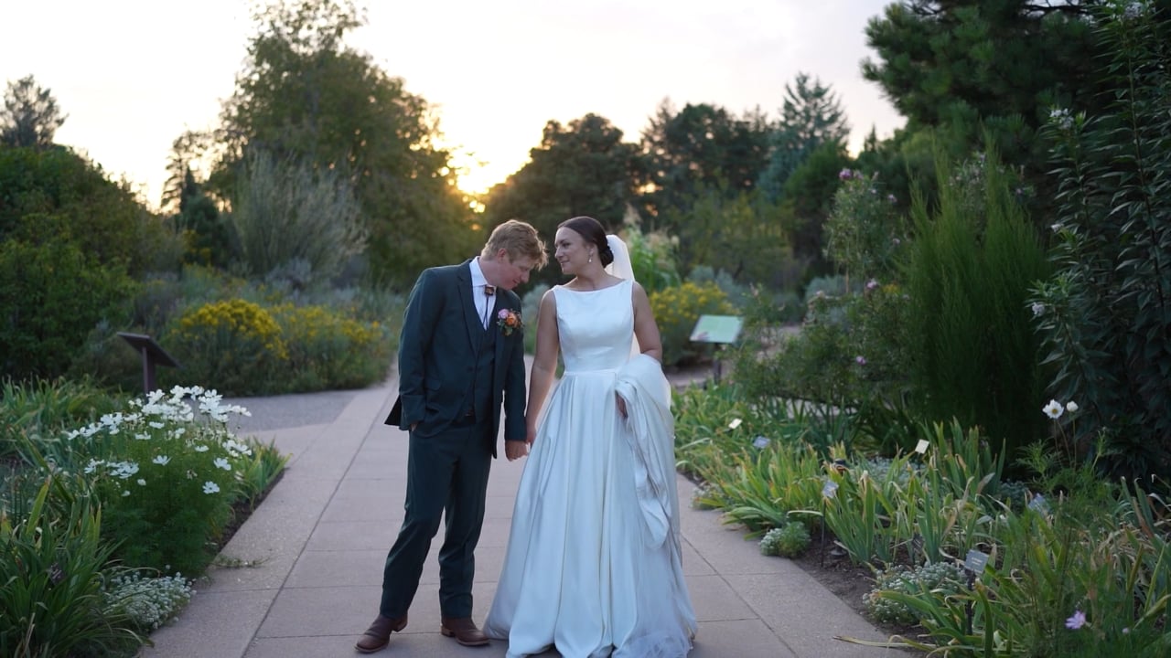 Claire and Will standing hand in hand on a garden path at the Denver Botanic Gardens as the sun sets behind the trees
