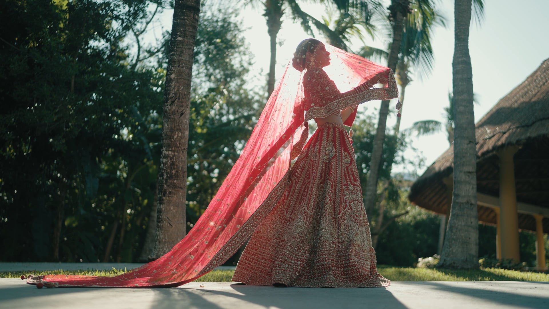 A bride in a red lehenga lifting her veil among palm trees &mdash; still from Richa & Shwetang's same day edit by Happy Soul Films