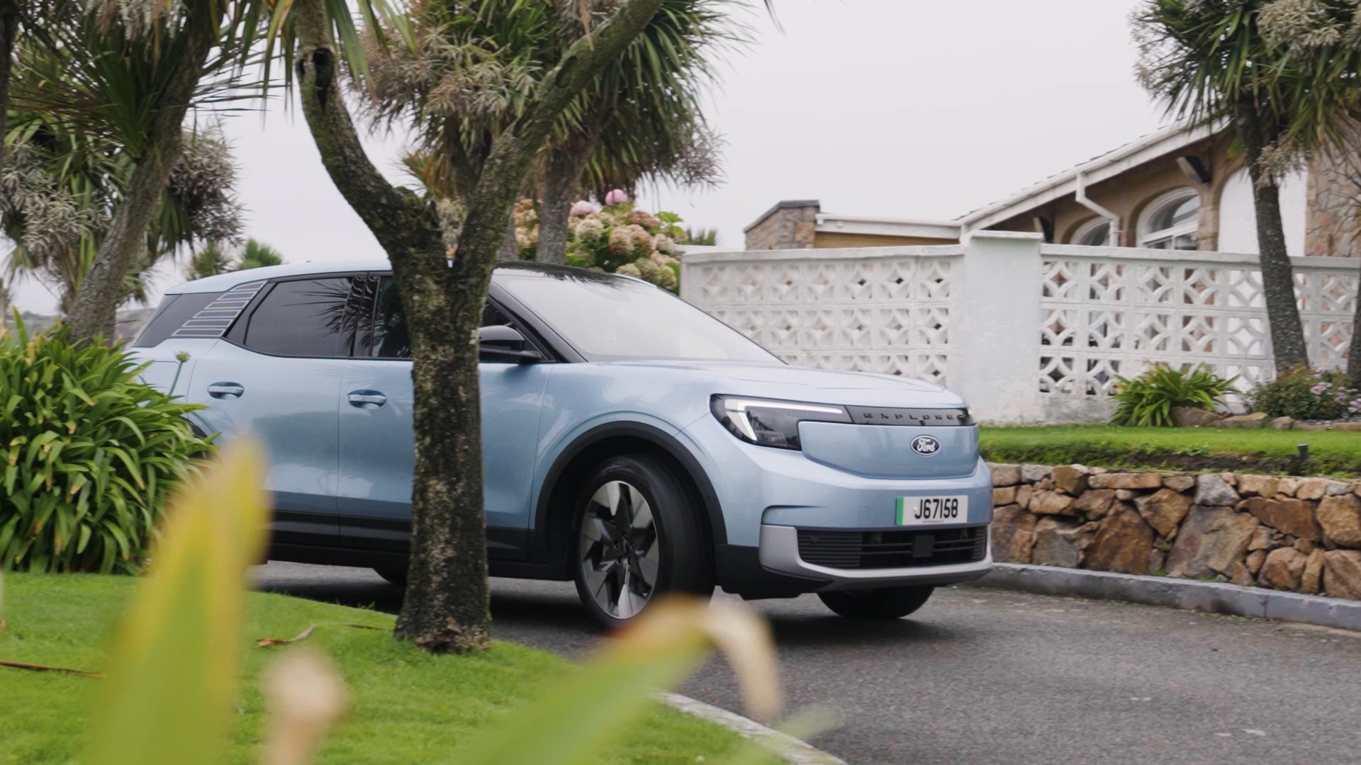 A light blue electice Ford Explorer pulling into a drivewhich has grass and palm trees either side. A house sits in the backgroud