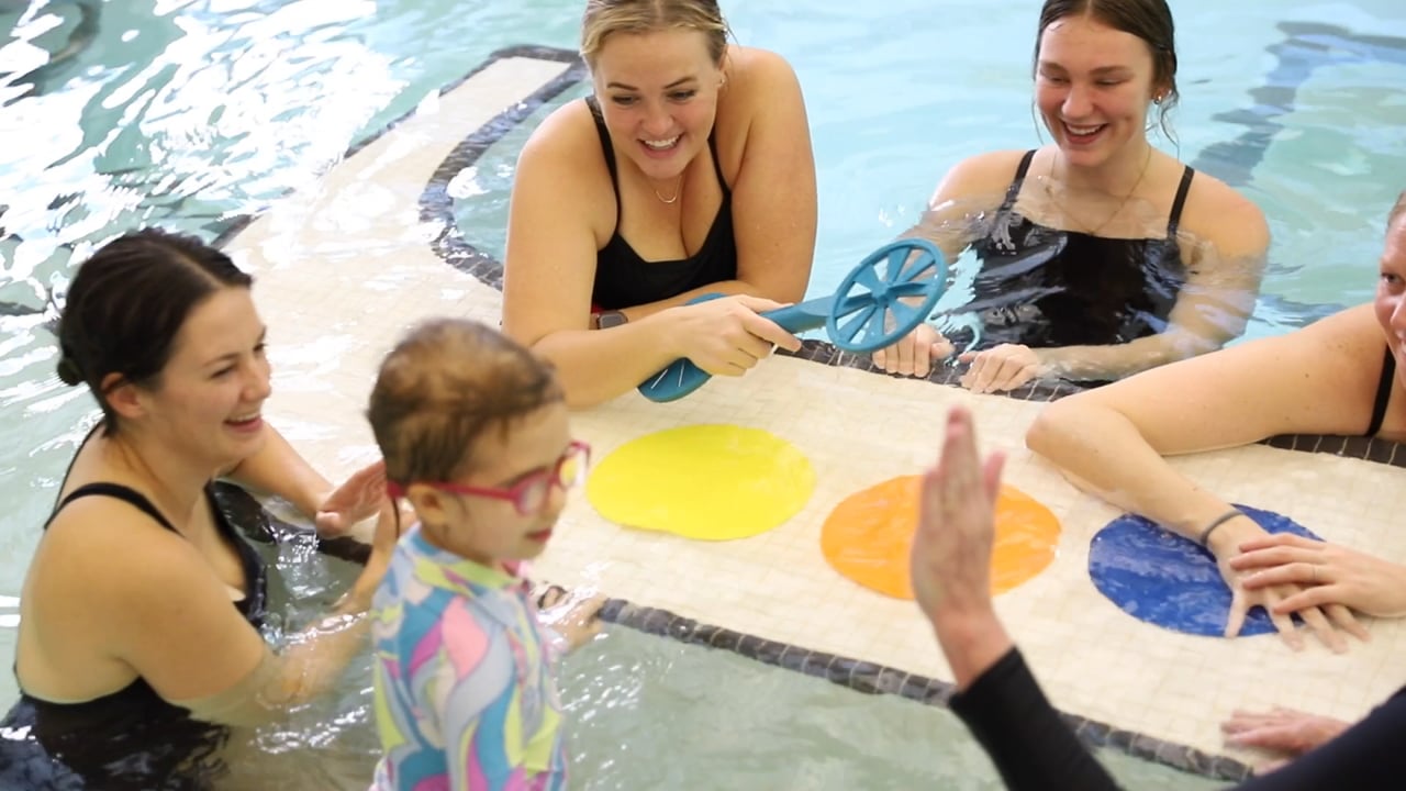 Smiling therapists in a pool with a young child at a floating mat of colored circles