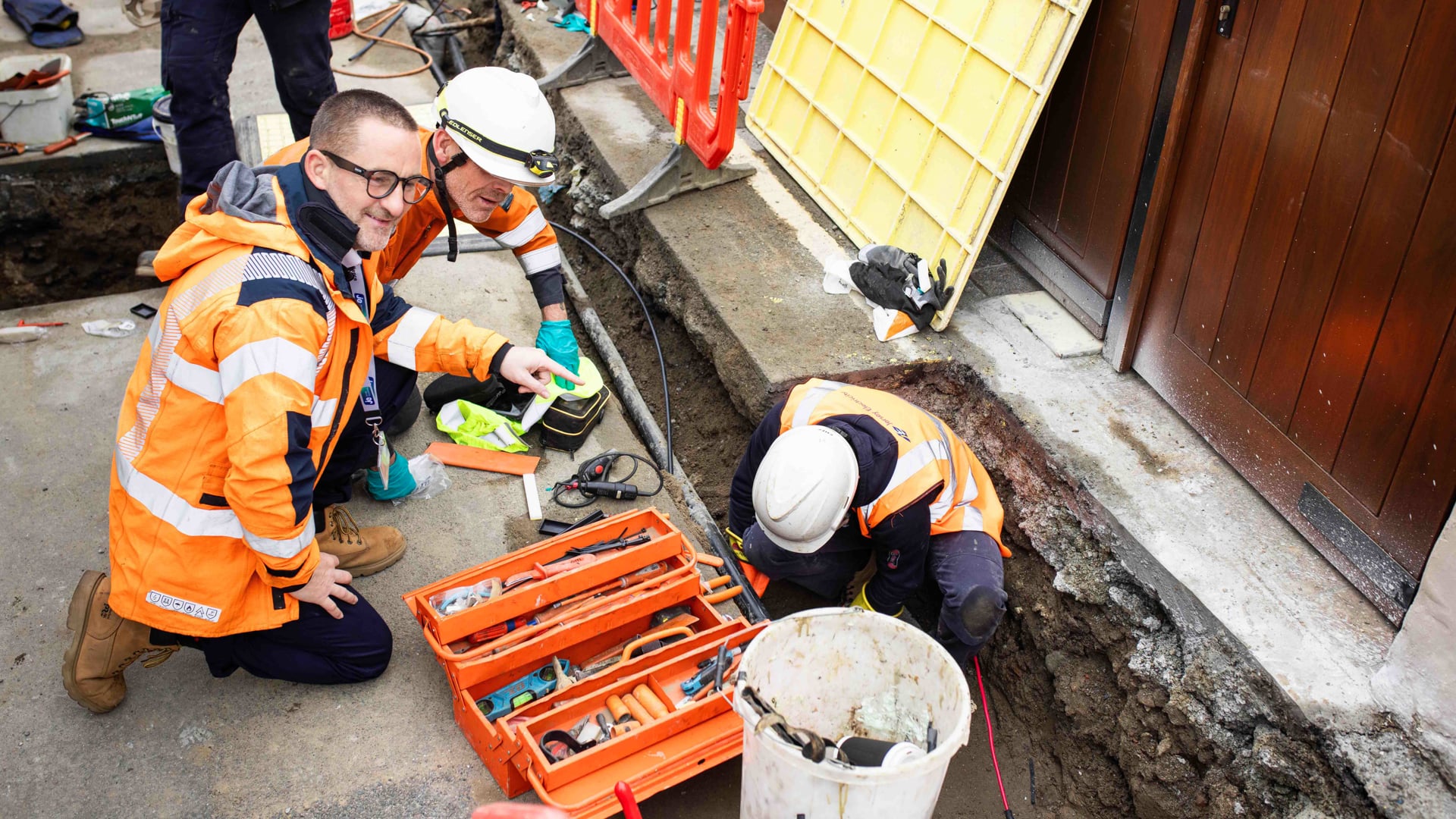 Two people wearing high vis jackets crouched down, looking into an open roadworks pit. There's a person in the pit wearing high vis and a hard hat