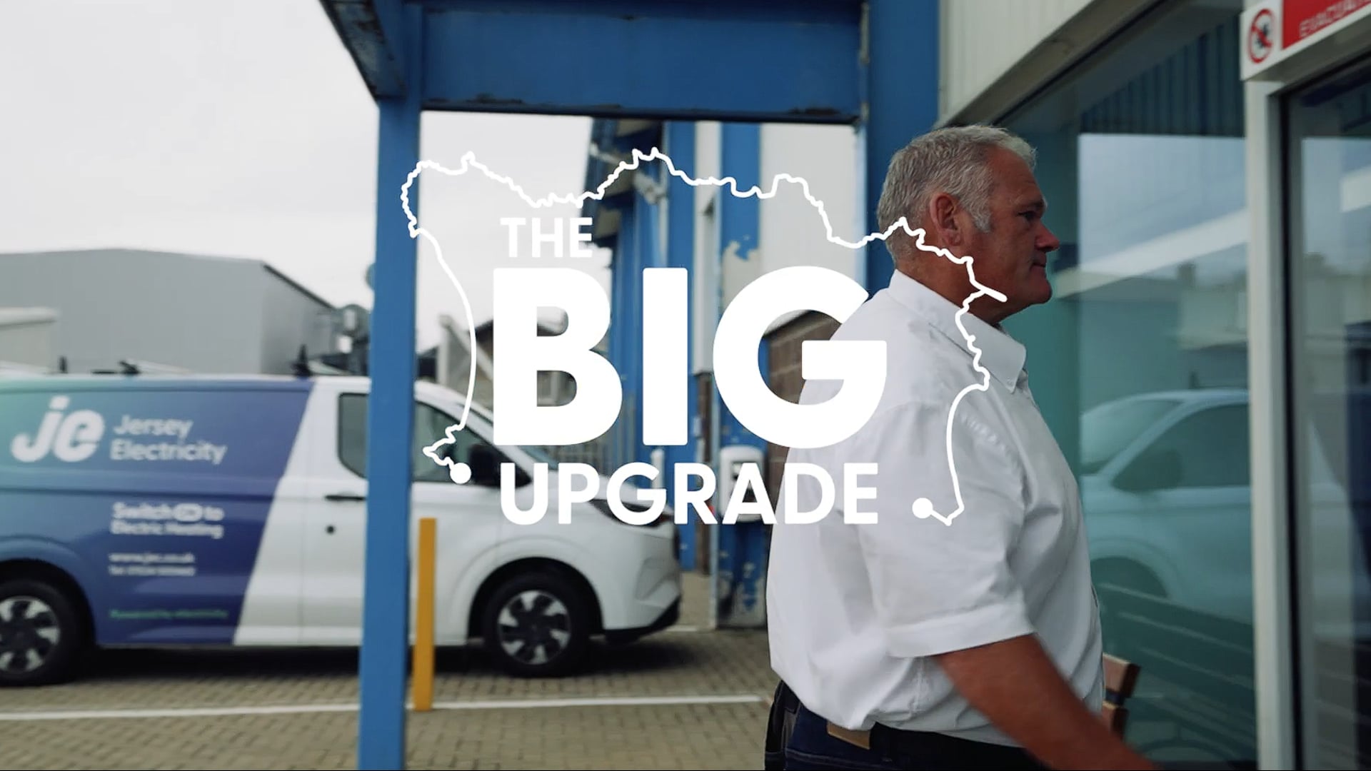 A person wearing a white shirt walking towards the power station. There's a Jersey Electricity van in the background and the title reading The BIG Upgrade in the foreground