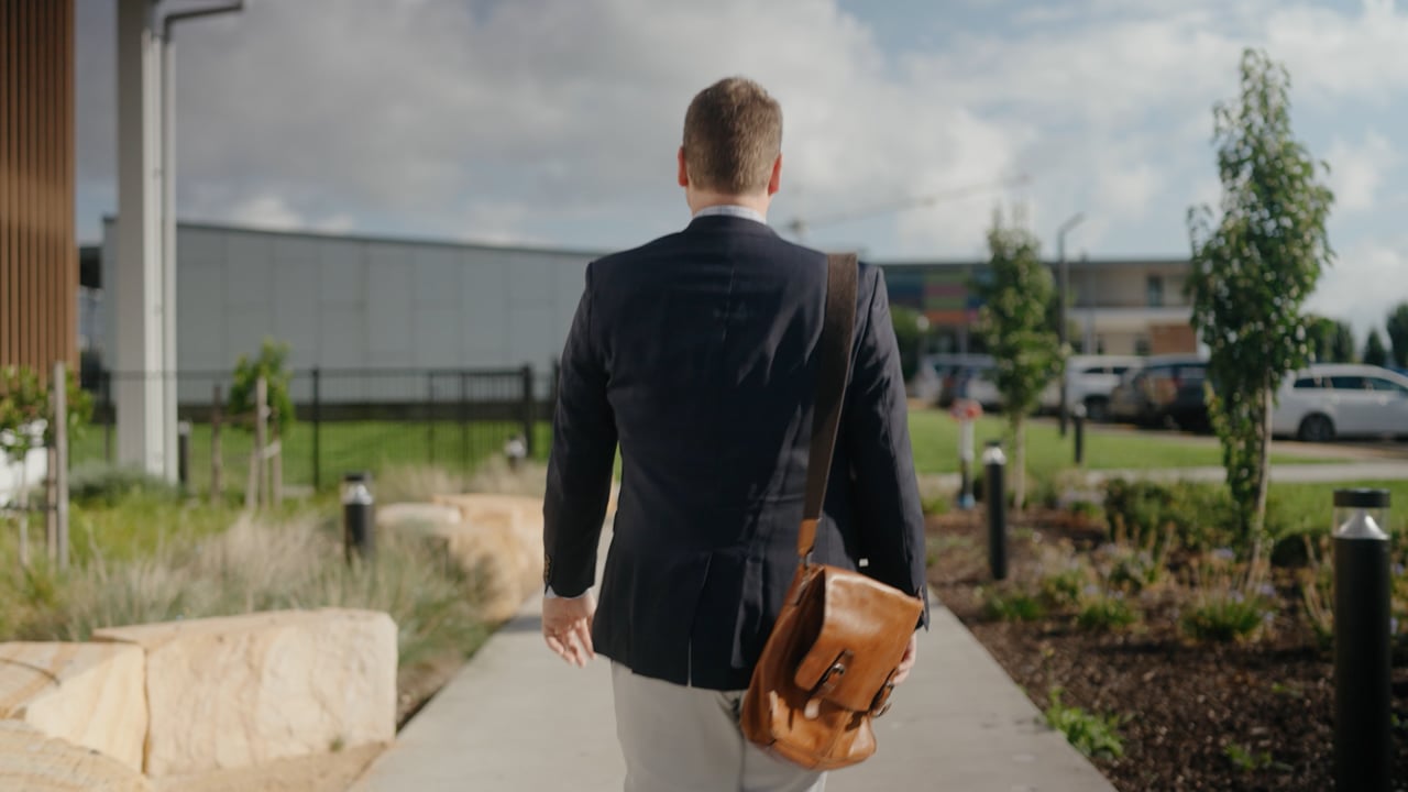 A man in a blazer carrying a leather satchel, walking a path through the Oran Park Anglican College grounds