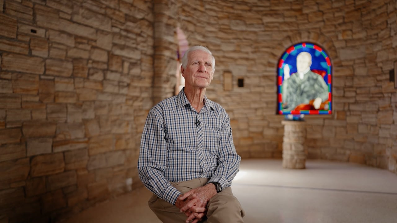 An older man seated in a sandstone chapel at Edmund Rice College, a stained glass window behind him