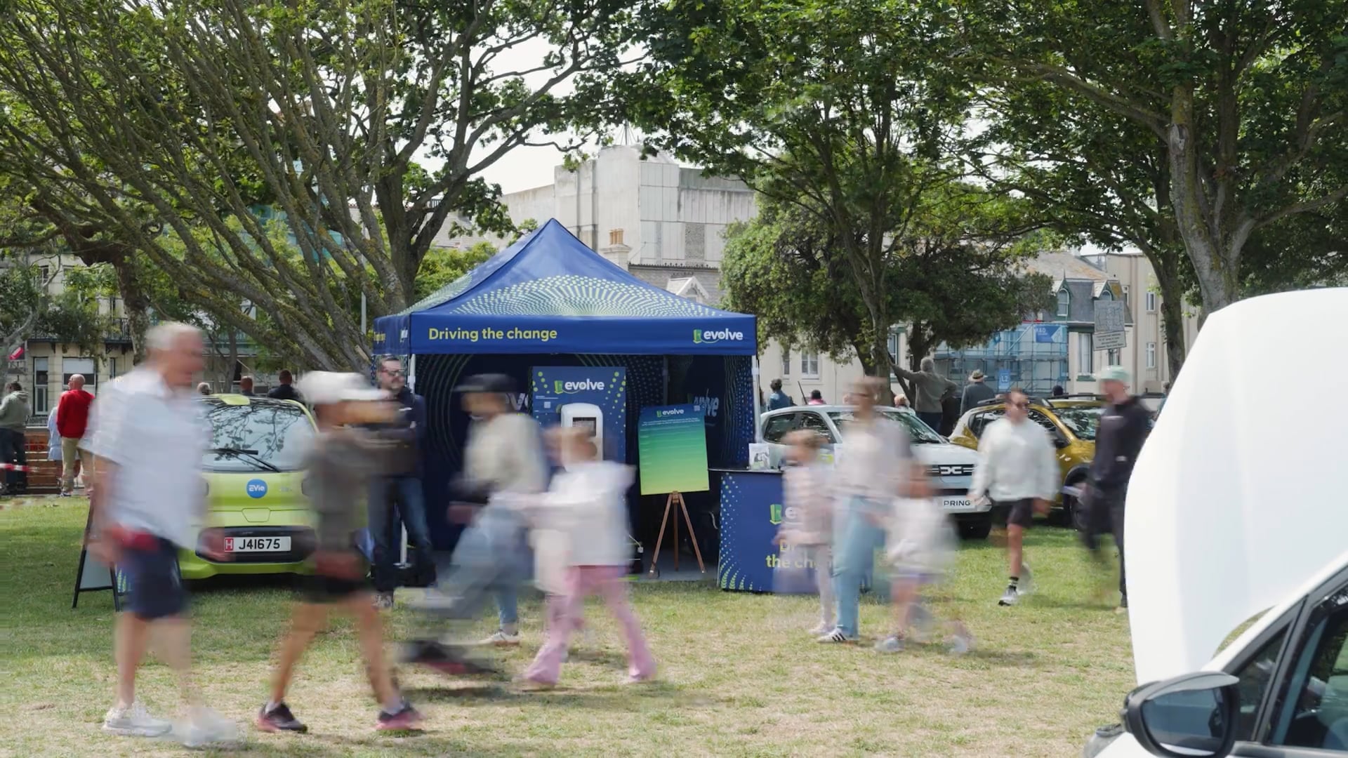 People walking past a blue "Driving the change" Evolve tent with electric cars on display in a park setting, with trees and buildings in the background.
