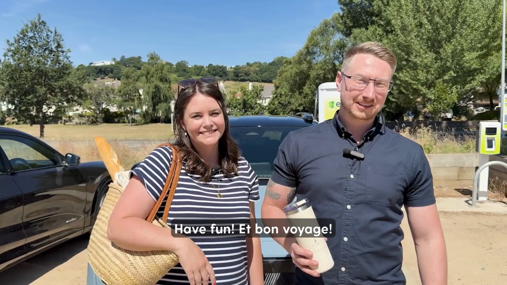 A woman with a striped shirt and a straw bag containing a baguette stands next to a black car, while a man in a collared shirt holding a reusable water bottle smiles. They are in an outdoor setting with greenery and what appears to be an electric vehicle charging station in the background. The text overlay reads "Have fun! Et bon voyage!".