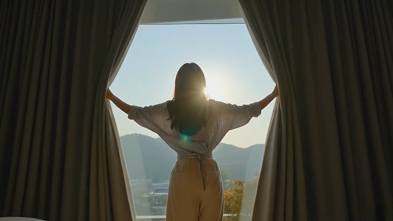 Woman opening hotel curtains to morning sunlight